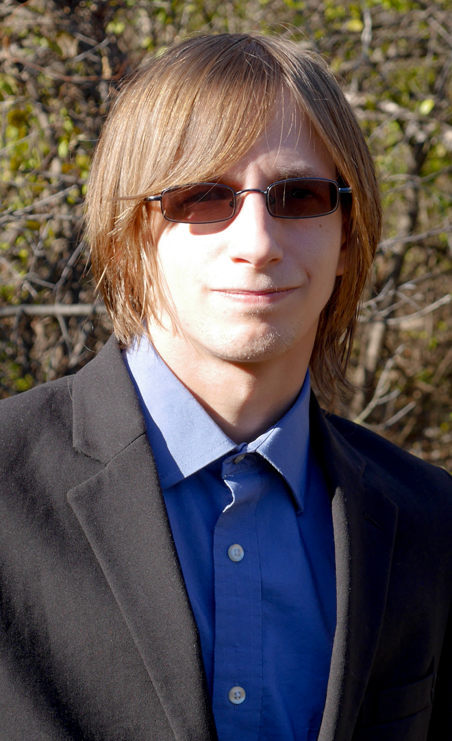 photo of a young man wearing glasses and a suit in front of blurred trees